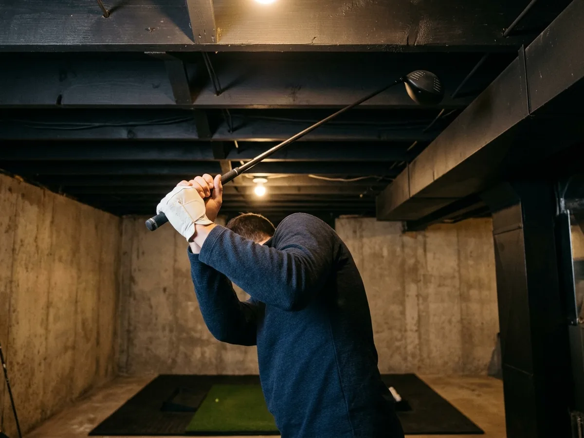 Golfer's gloved hands at the top of a driver backswing under a basement ceiling with open joists painted black